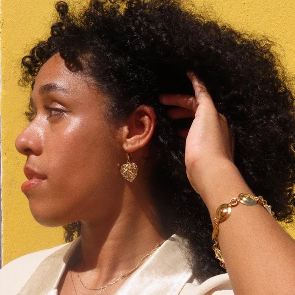 Perfil de modelo con pantalla dorada de corazón y pulsera de eslabones dorados contra pared amarilla