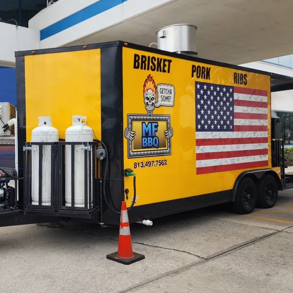 El trailer amarillo de MF BBQ con la bandera americana y las palabras Brisket, Pork, Ribs