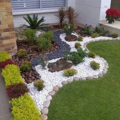Jardín con piedra blanca, borde de río y arbustos de colores contra casa de ladrillo