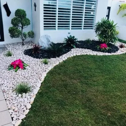 Jardín de piedra blanca con mantillo negro, topiario y bromelias frente a casa blanca en Puerto Rico