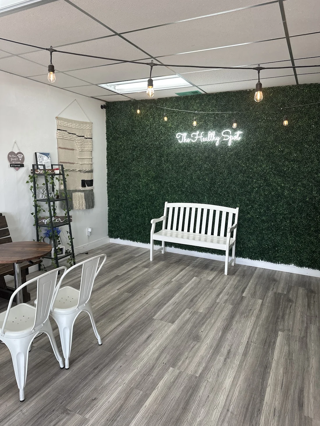 Salón del local con pared verde de boxwood, letrero de neón The Healthy Spot, banco blanco y bombillas colgando