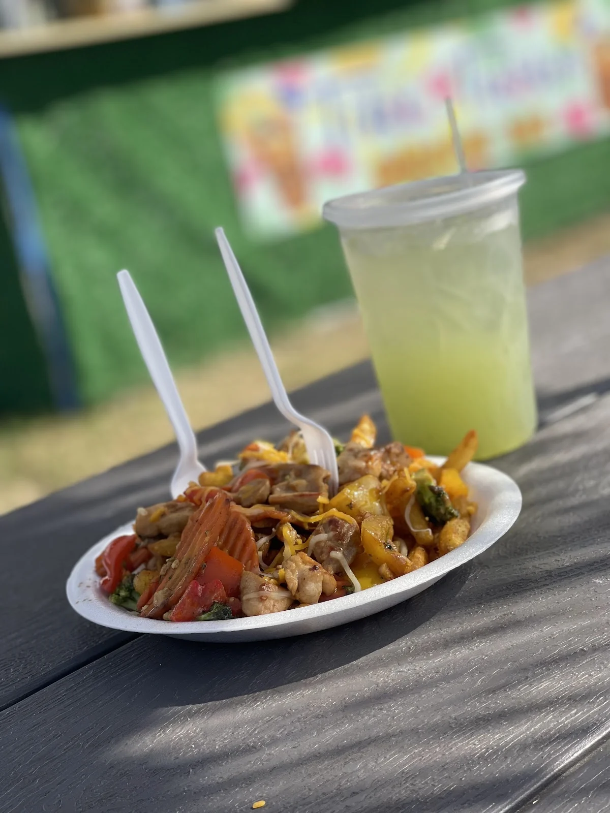 Vaso de limonada fresca de 24oz junto a un plato de stir fry sobre la mesa de picnic, con el letrero de Tiki Turbo detrás