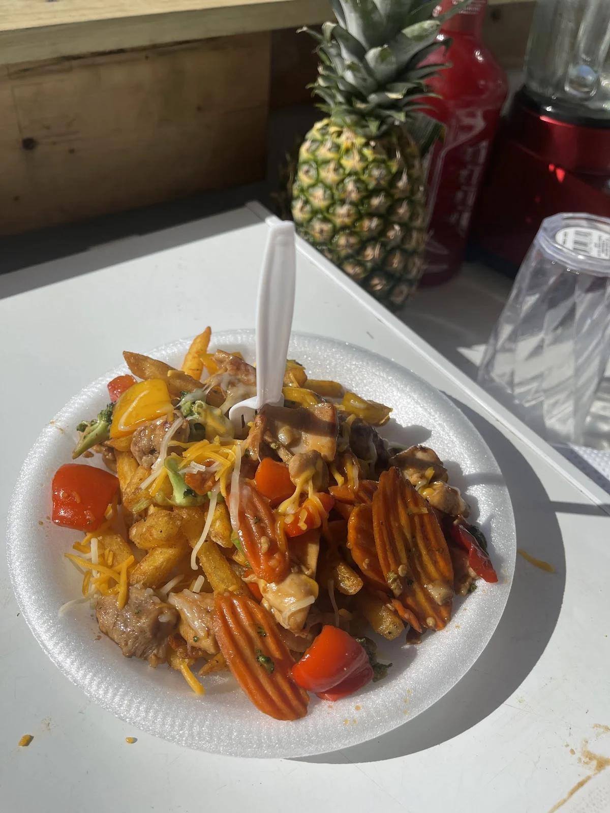 Plato de stir fry con carne, pimientos, zanahoria, brócoli y queso, junto a una piña entera y sriracha