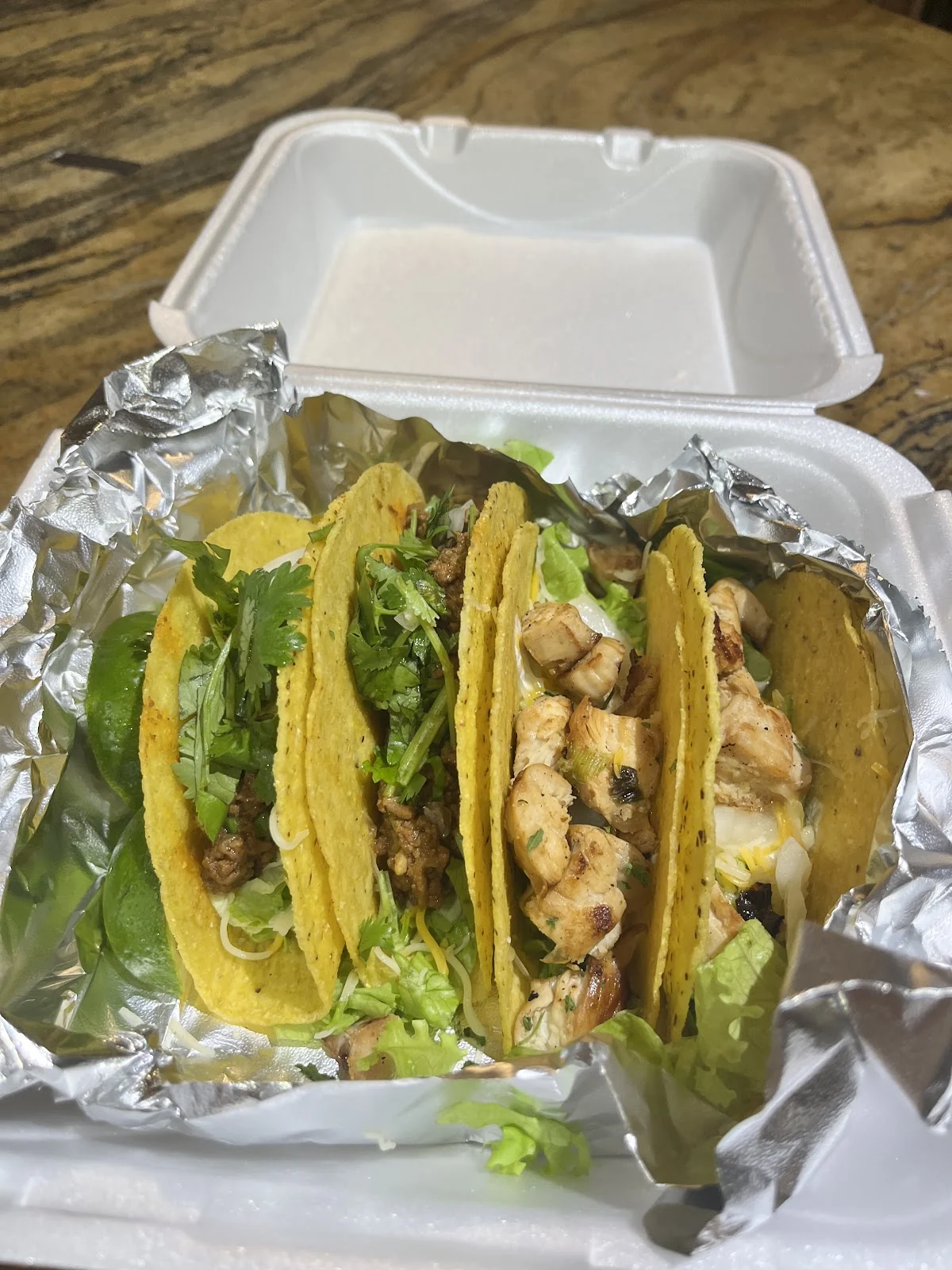 Cuatro tacos con carne molida y pollo, cilantro, lechuga y queso, servidos en papel aluminio